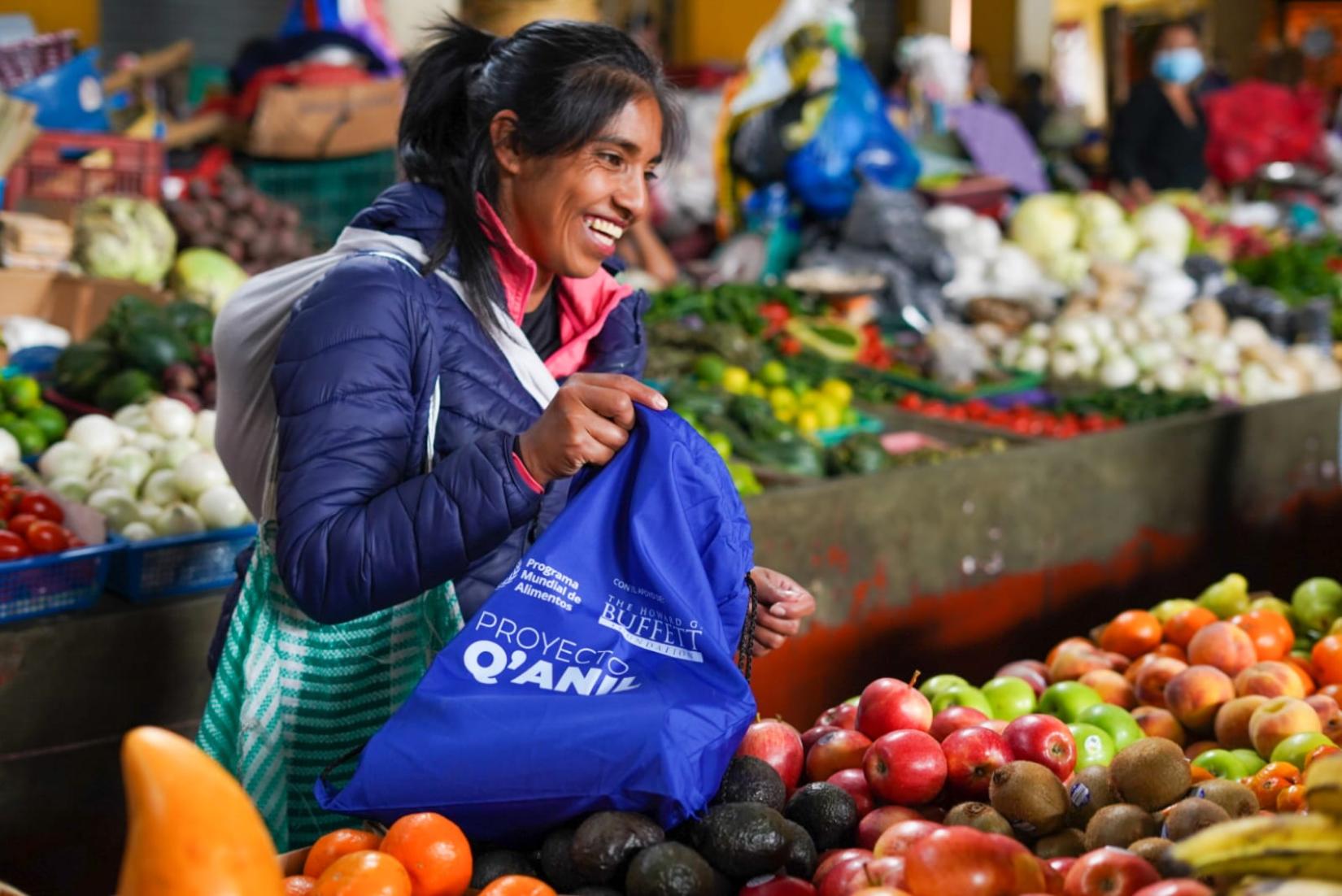 Mujer en una venta de frutas, sonriendo, llena bolsa con distintivo del proyecto Q'anil, con los productos expuestos al público