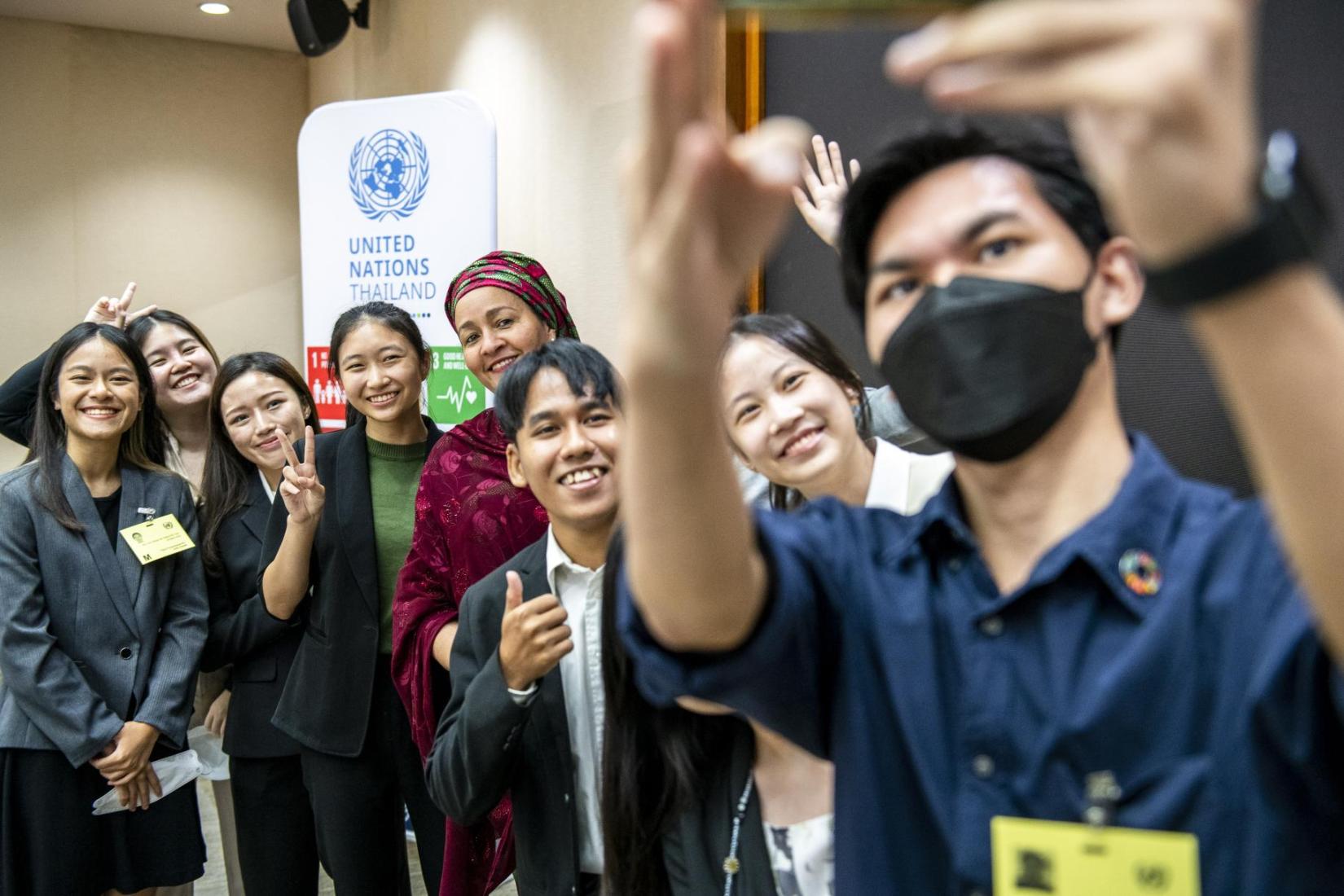 Imagen grupo de jóvenes se toman una selfie con una mujer con pañuelo verde y rojo frente a una pancarta de la ONU Pie de foto: En Tailandia, el consejo asesor de jóvenes para los ODS está ayudando a crear conciencia a través de plataformas digitales como Instagram.